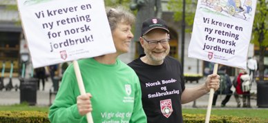 Kvinne i grønn genser og mann i svart t-skjorte og caps smiler. De holder hver sin plakat hvor det står "vi krever en ny retning for norsk jordbruk". De er på markering foran Stortinget.  - Foto: Nora May Engeseth/NBS