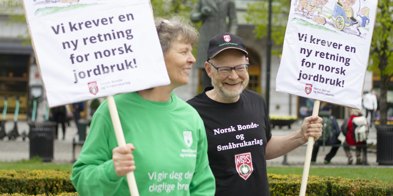 Kvinne i grønn genser og mann i svart t-skjorte og caps smiler. De holder hver sin plakat hvor det står "vi krever en ny retning for norsk jordbruk". De er på markering foran Stortinget.  - Foto: Nora May Engeseth/NBS