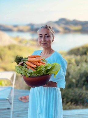 Woman in blue dress holds a bowl of lettuce and carrots she has grown herself.  In the background we see a coastal landscape.