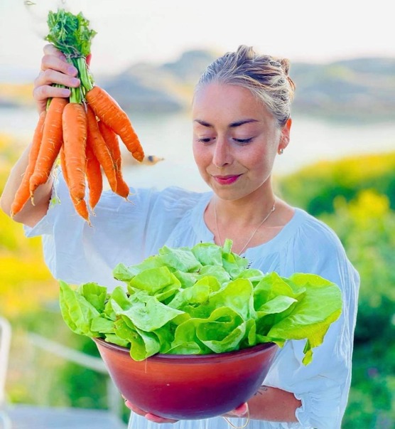 A woman in a blue dress holds a handful of carrots in one hand, and an earthenware bowl with salad in the other.  She looks at the salad bowl happily.
