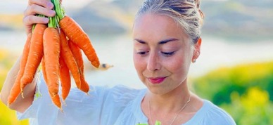 La giovane, vestita di blu, tiene in mano un fagotto di carote.  D'altra parte ha una grande ciotola di insalata fresca.  La donna guarda l'insalata.  - Foto: Gilberto Debelian La giovane, vestita di blu, tiene in mano un fagotto di carote.  D'altra parte ha una grande ciotola di insalata fresca.  La donna guarda l'insalata.  - Foto: Gilberto Debelian